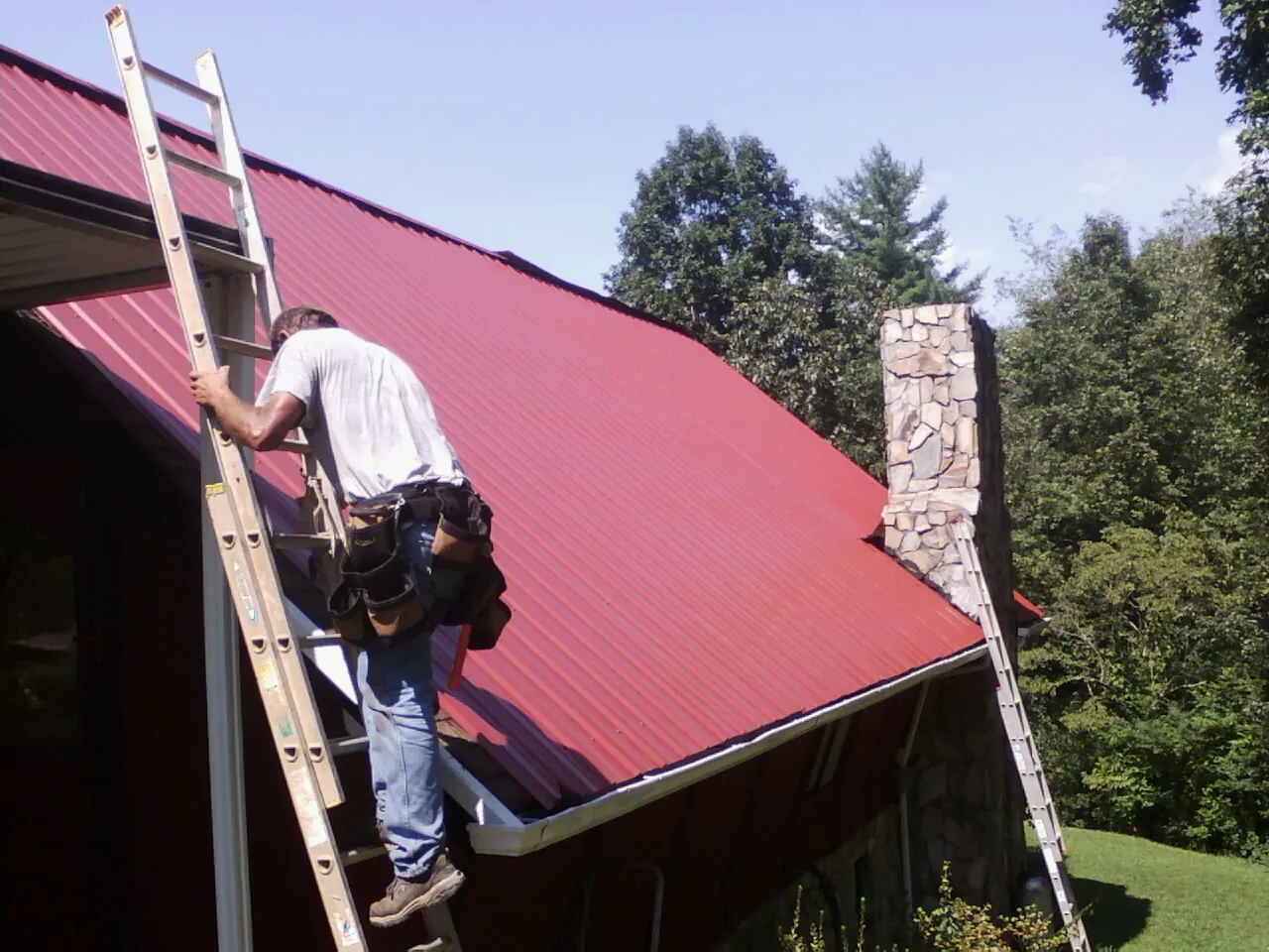 Roofer on a ladder beside a red metal roof and stone chimney — Soffit & Fascia Repair by Roofing Brothers in Carolina Beach